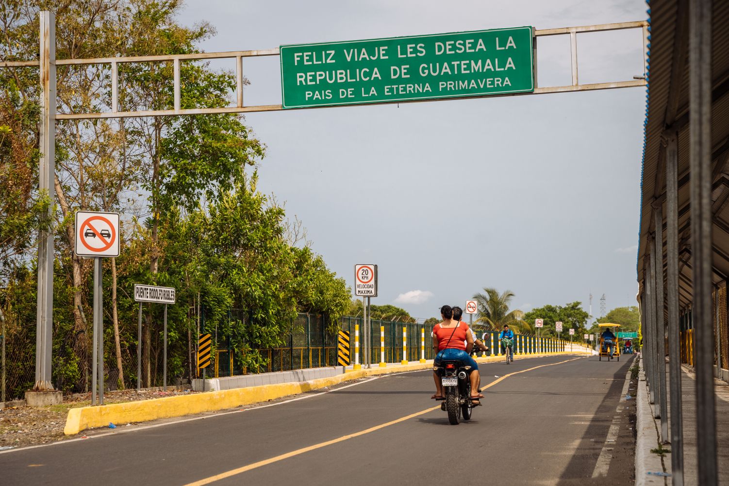 The backs of two people on a motorbike; above them a sign notes the Guatemala border