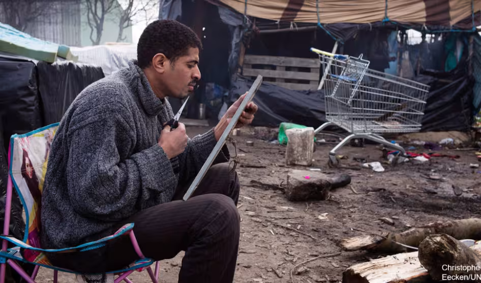 An asylum seeker trims his beard in a mirror on his lap