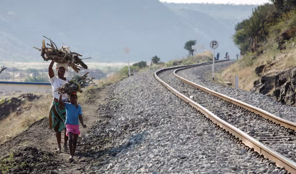 A woman and child walk by a railroad with large piles of wood on their heads.