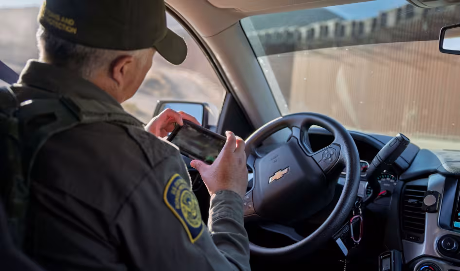 A male border patrol agent in his vehicle looks at a mobile device.