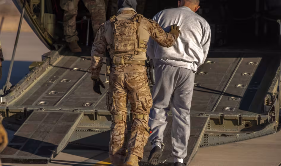 A person in fatigues pushes a detained man into the back of an airplane.