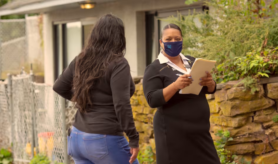 Image of masked woman with a clipboard speaking with another woman outside her home.