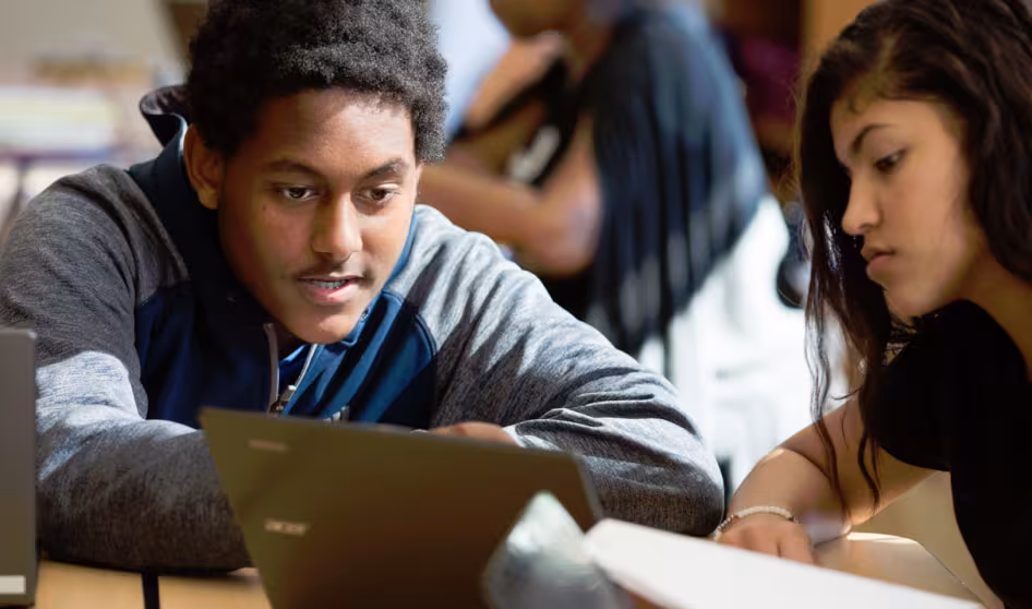 Image of two students in a classroom, one with a laptop