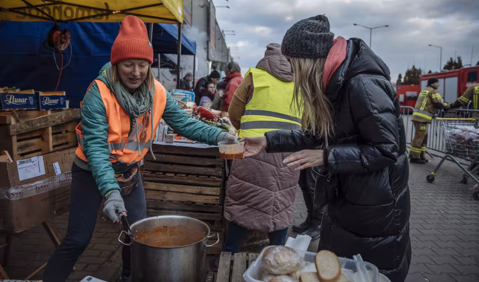 A woman in a winter coat receives food from a woman in an orange safety vest at a train station in P
