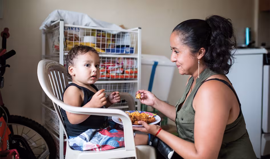 A woman feeds a young child sitting on a white plastic chair 
