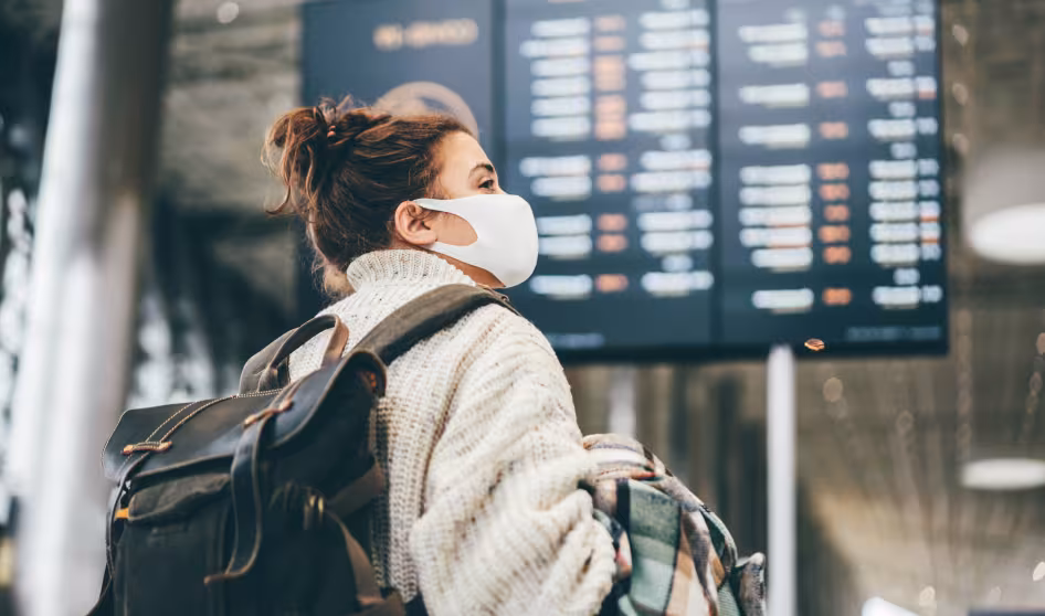 Woman wearing a white sweater and face mask and carrying luggage looks at airport information board