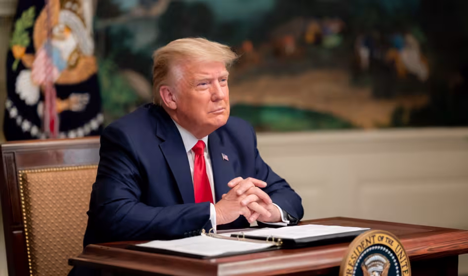 President Trump sits with his hands clasped at a desk with papers and a pen in front of him