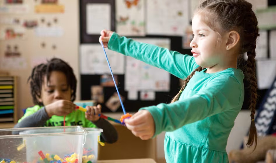 Two preschool students thread beads on a string in a classroom