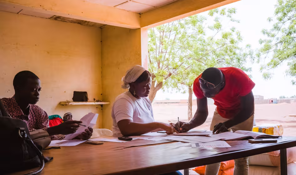 Two people write on papers while another sits nearby. Migrants returning to Burkina Faso receive food and non-food items as reintegration assistance.