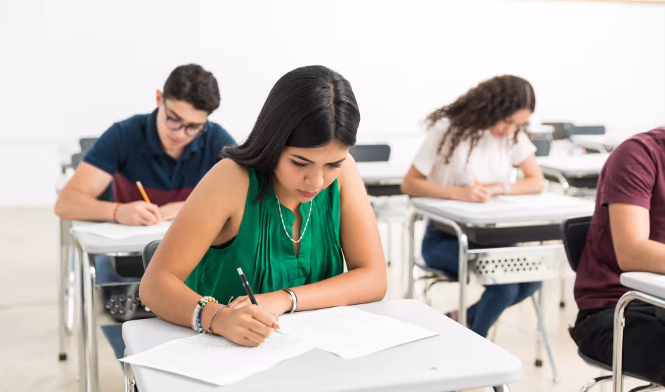 A Latina high school student in a green shirt sits at a desk and takes a test; other students do the same in the background