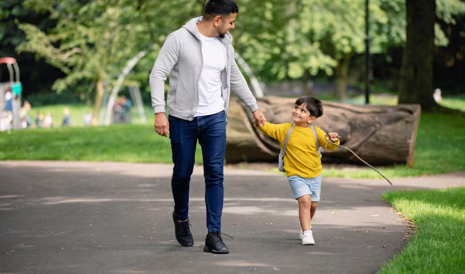 A young boy holds hands with his dad as they walk in a park