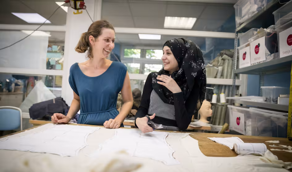 German seamstress works with a trainee Syrian refugee at a fashion workshop