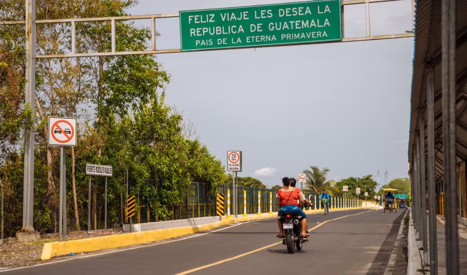 The backs of two people on a motorbike; above them a sign notes the Guatemala border
