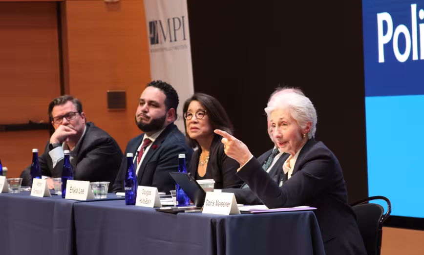 A woman, Doris Meissner, speaks at a panel discussion and points to the audience
