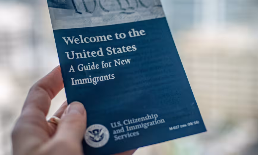 A person holds a document reading "Welcome to the United States: A Guide for Immigrants"