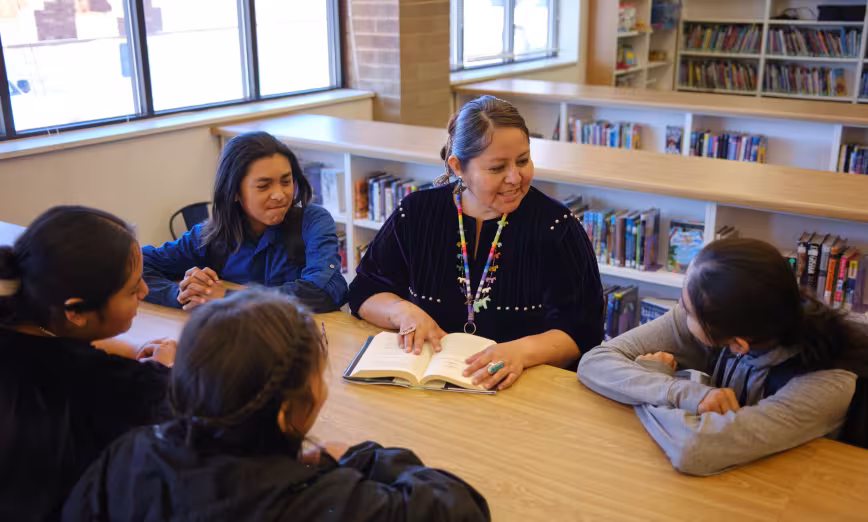  A high school teacher and students sit around a table in a school library
