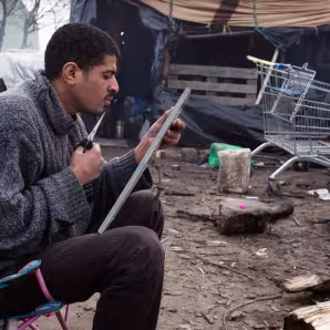 An asylum seeker trims his beard in a mirror on his lap