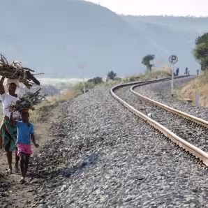 A woman and child walk by a railroad with large piles of wood on their heads.