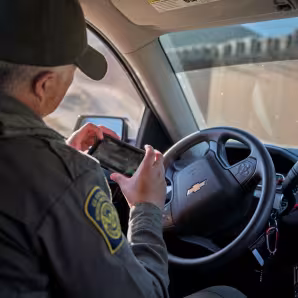 A male border patrol agent in his vehicle looks at a mobile device.