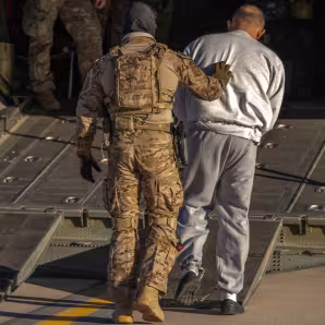 A person in fatigues pushes a detained man into the back of an airplane.