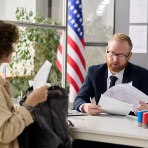 A man in a suit reviews a woman's paperwork, while the U.S. flag stands behind him