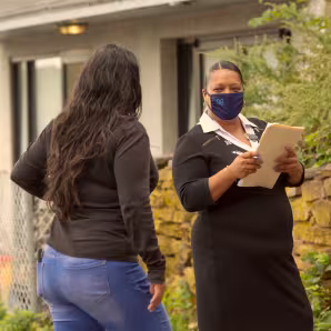 Image of masked woman with a clipboard speaking with another woman outside her home.