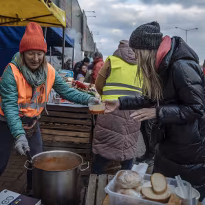 A woman in a winter coat receives food from a woman in an orange safety vest at a train station in P