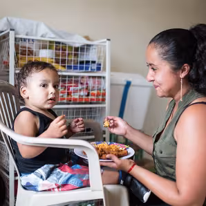 A woman feeds a young child sitting on a white plastic chair 