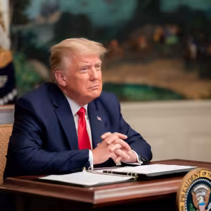 President Trump sits with his hands clasped at a desk with papers and a pen in front of him