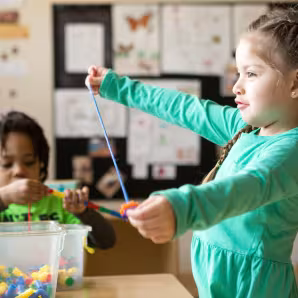 Two preschool students thread beads on a string in a classroom