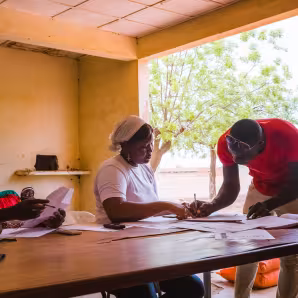 Two people write on papers while another sits nearby. Migrants returning to Burkina Faso receive food and non-food items as reintegration assistance.