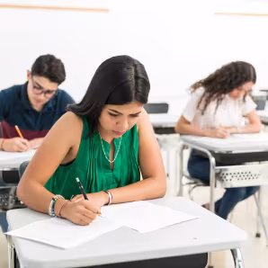 A Latina high school student in a green shirt sits at a desk and takes a test; other students do the same in the background