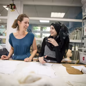 German seamstress works with a trainee Syrian refugee at a fashion workshop
