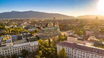 Aerial golden-hour view of Sofia, with Alexander Nevsky Cathedral's gold and green copper domes rising above the city and Vitosha Mountain beyond