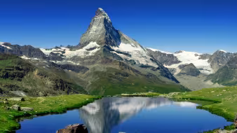 The Matterhorn reflected in Riffelsee lake near Zermatt, with green alpine meadows, rocky terrain, and snow-capped peaks under a blue sky