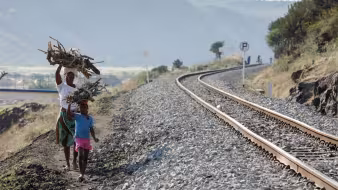 A woman and child walk by a railroad with large piles of wood on their heads.