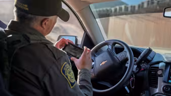 A male border patrol agent in his vehicle looks at a mobile device.