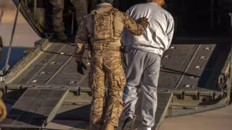 A person in fatigues pushes a detained man into the back of an airplane.