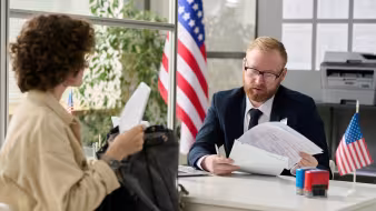 A man in a suit reviews a woman's paperwork, while the U.S. flag stands behind him