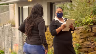 Image of masked woman with a clipboard speaking with another woman outside her home.