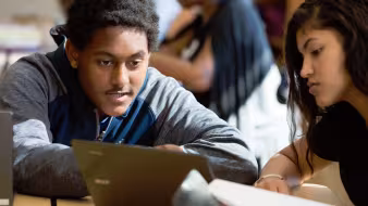 Image of two students in a classroom, one with a laptop