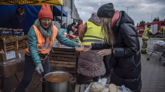 A woman in a winter coat receives food from a woman in an orange safety vest at a train station in P