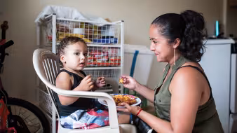 A woman feeds a young child sitting on a white plastic chair 