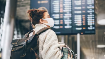 Woman wearing a white sweater and face mask and carrying luggage looks at airport information board