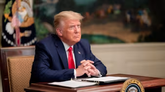 President Trump sits with his hands clasped at a desk with papers and a pen in front of him