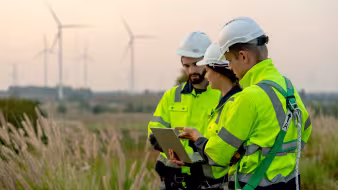 Three technicians stand in a field in front of wind turbines.