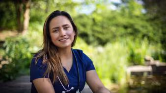 A young female nurse wearing medical scrubs and a stethoscope sits on a park bench