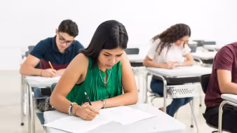 A Latina high school student in a green shirt sits at a desk and takes a test; other students do the same in the background