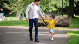 A young boy holds hands with his dad as they walk in a park