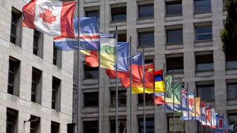 At least 20 flags of different countries fly in the wind outside of two large buildings